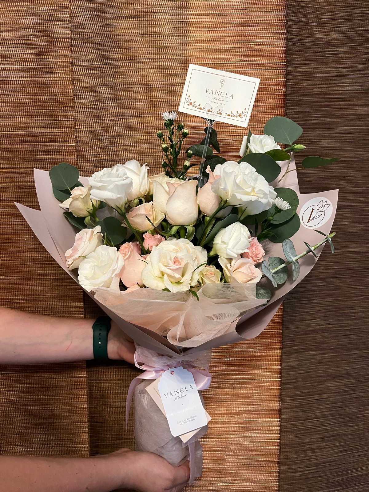 Person holding a bouquet of white and pink roses with eucalyptus leaves wrapped in pink tissue paper against a brick wall