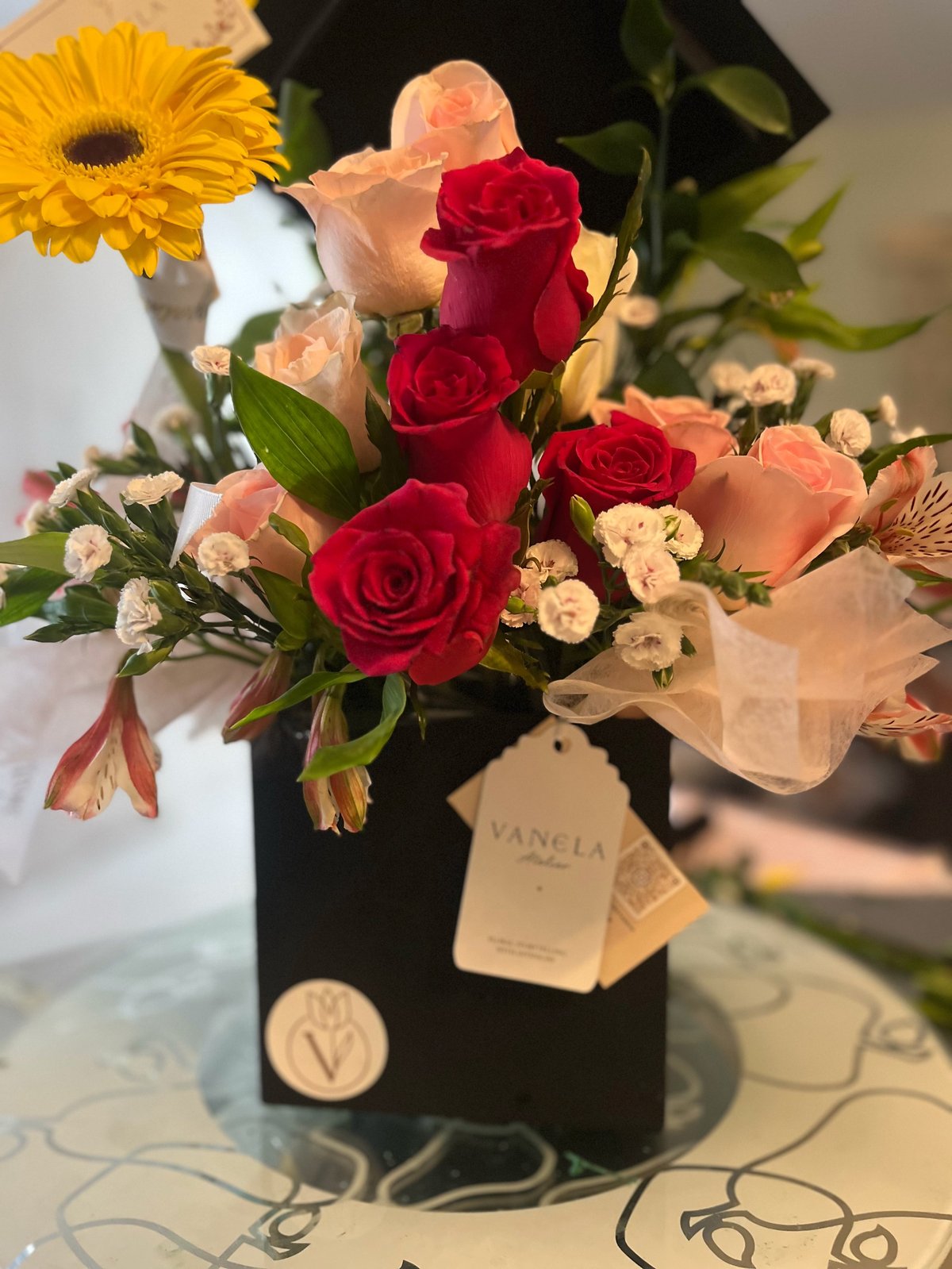 Floral arrangement in black vase with red roses, peach roses, yellow gerbera daisy, and small white flowers with sheer ribbon bow
