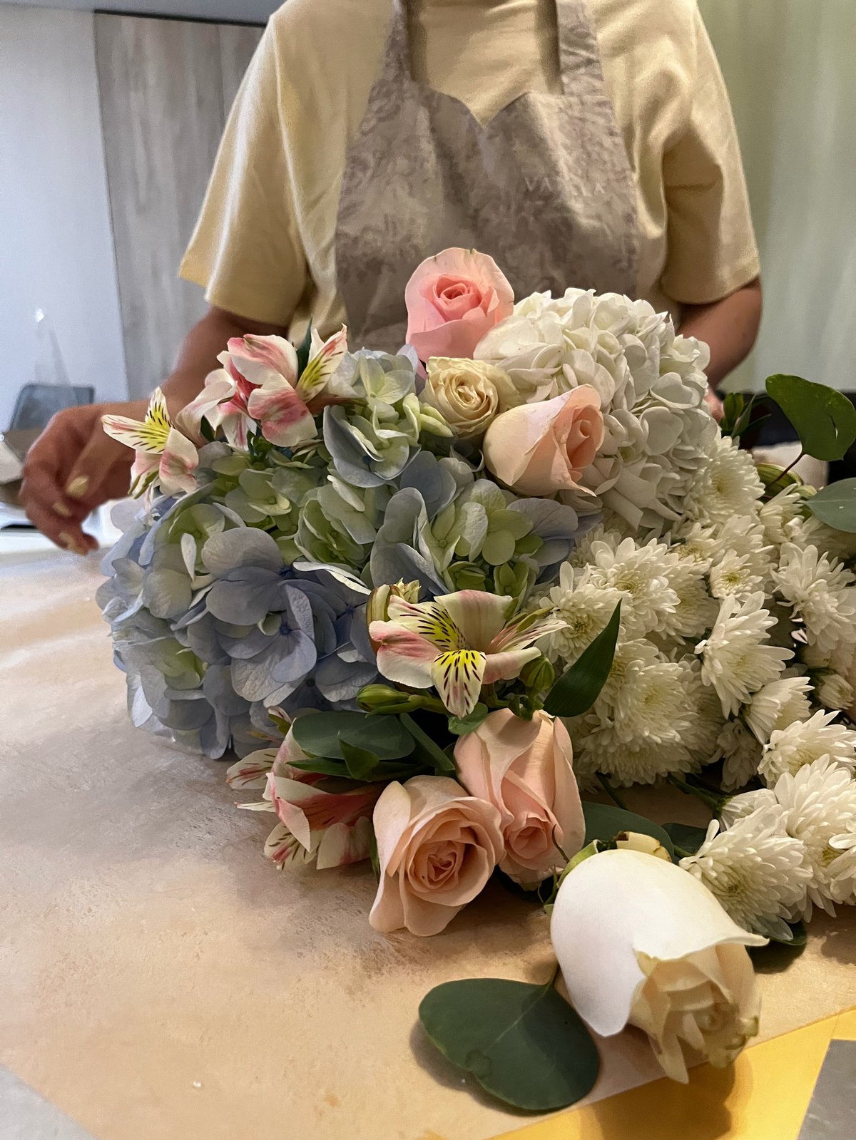 Person in yellow shirt arranging a bridal bouquet with pink roses, cream carnations, blue hydrangeas, and white flowers on a table.