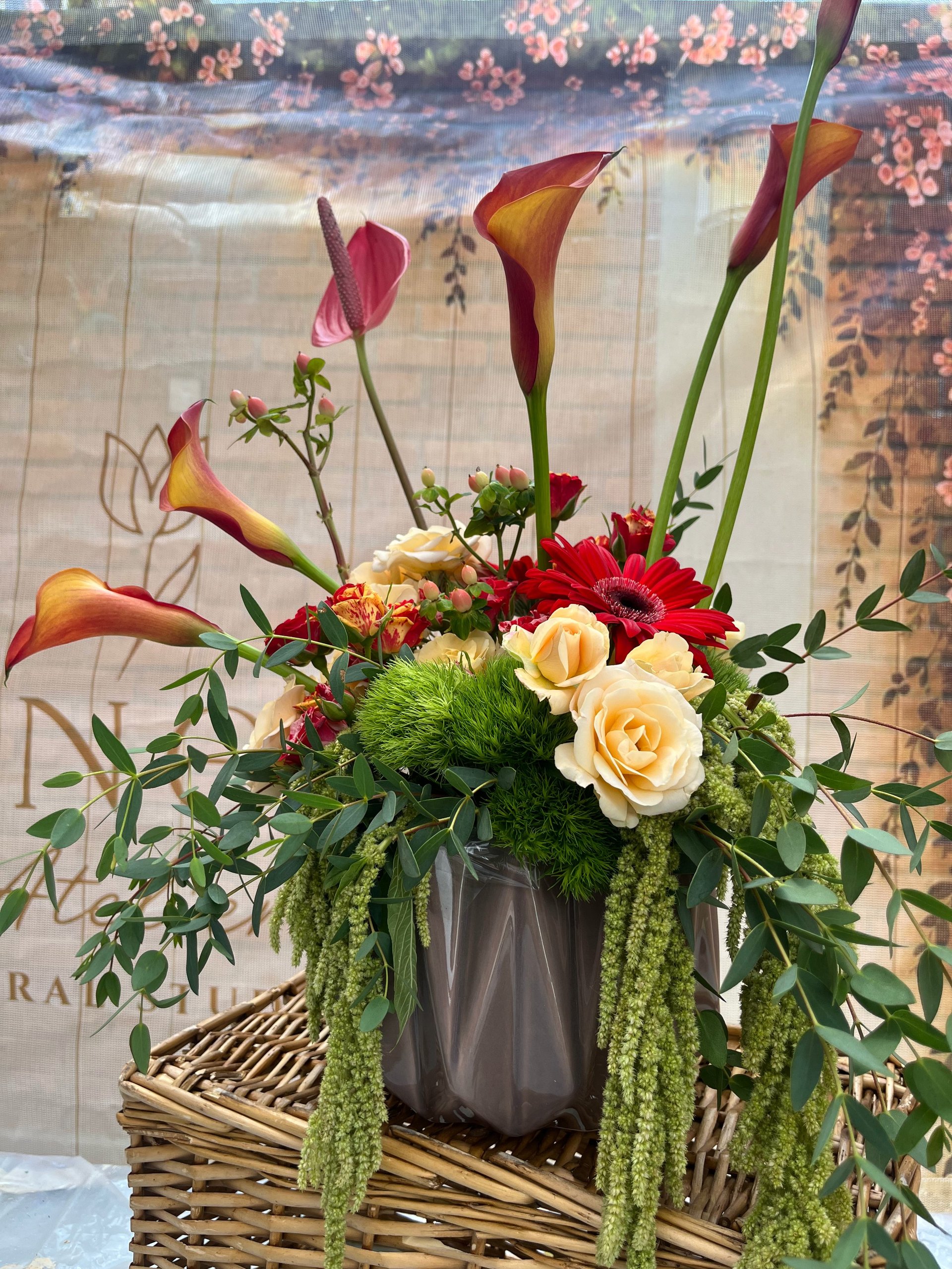 Colorful floral arrangement in a dark vase with calla lilies, roses, red gerberas, and trailing greenery displayed on a woven basket