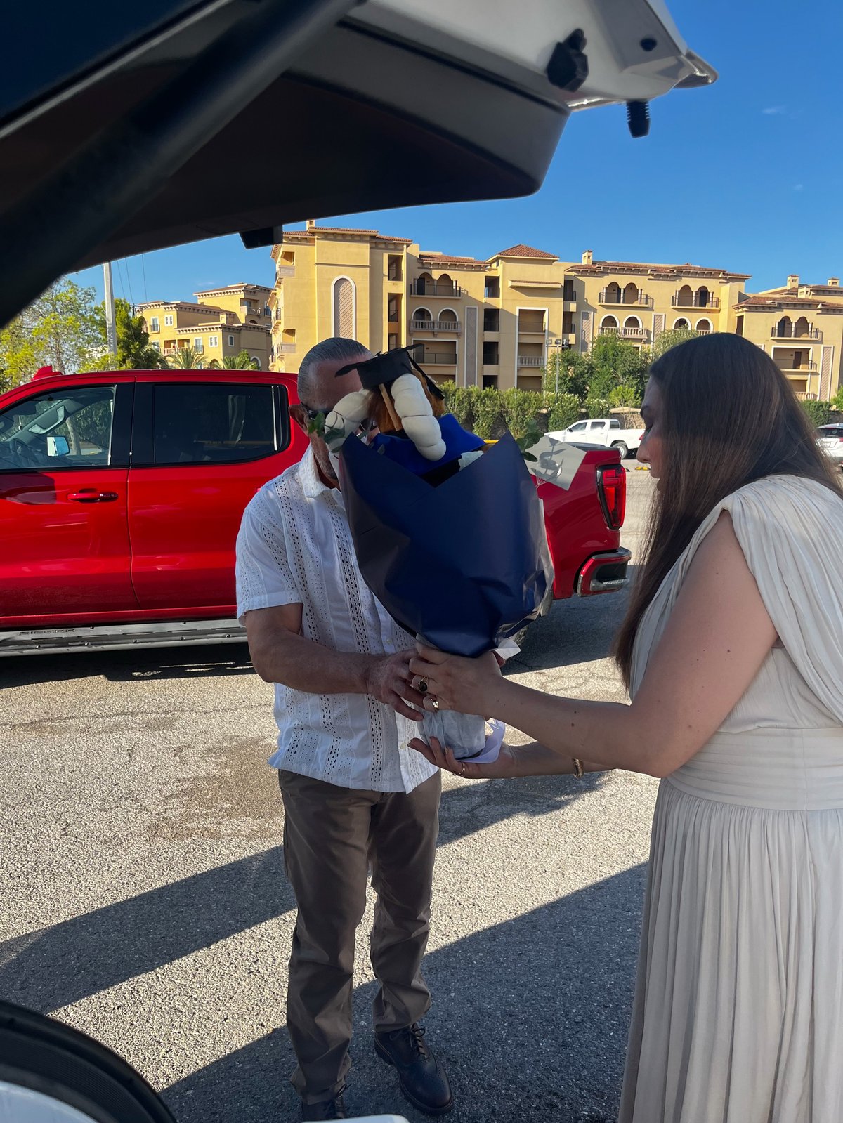 Man and woman stand between red cars, man holding blue fabric overhead in sunny parking lot with yellow building in background