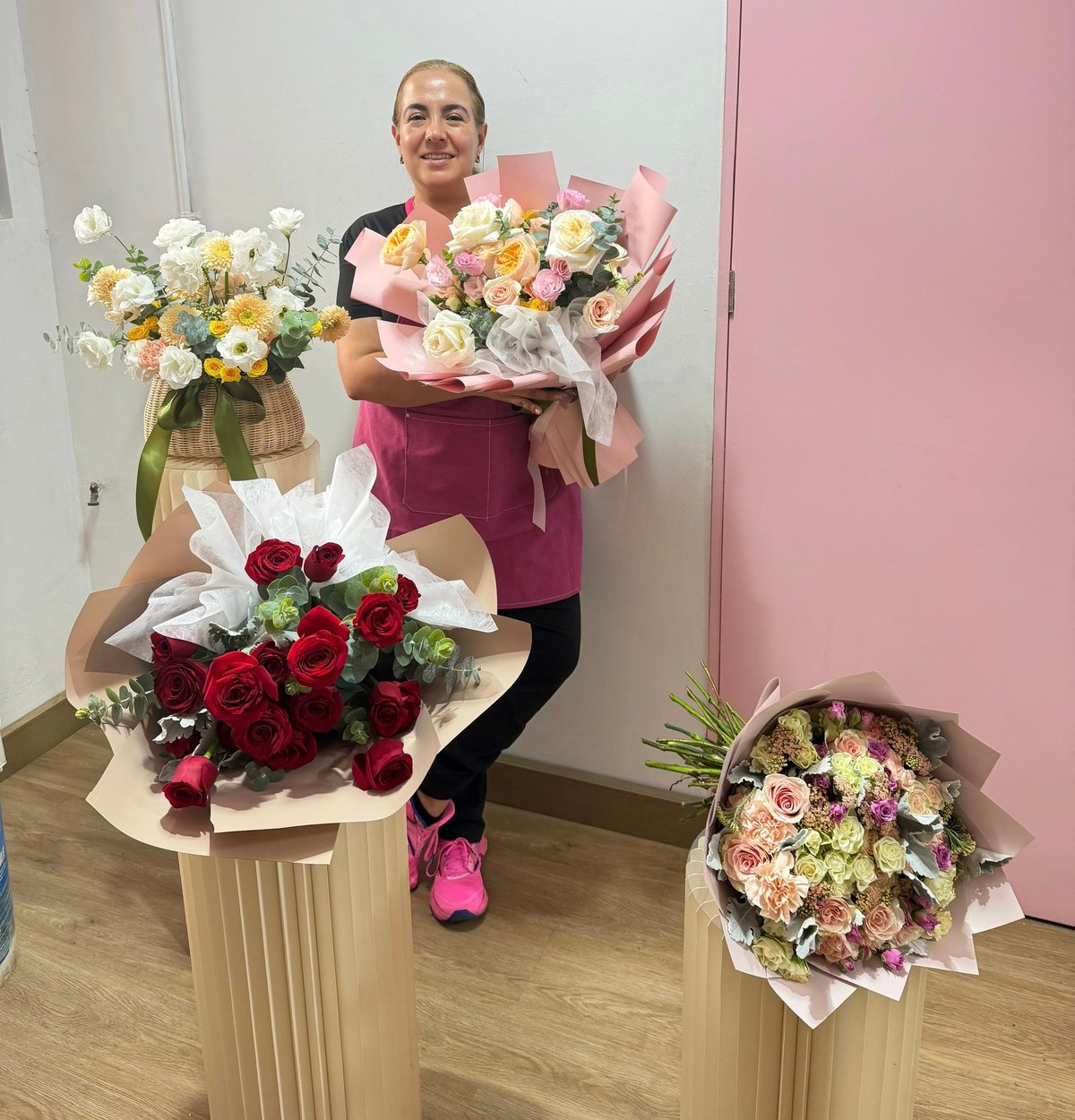 Florist holding bouquets of pink and yellow flowers, surrounded by arrangements including red roses on pedestals in a shop.
