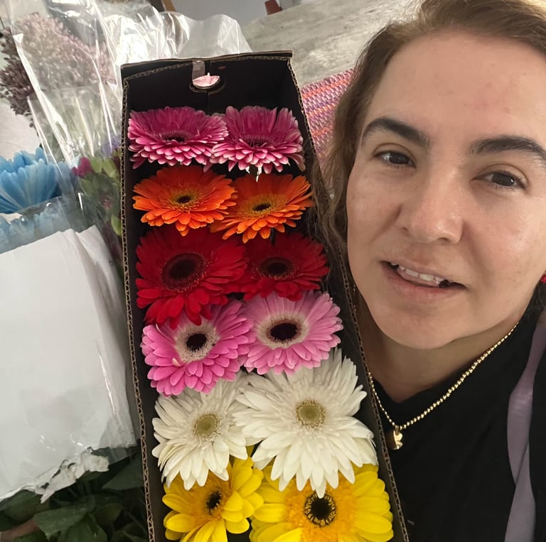 Woman smiling next to colorful gerbera daisies in a black gift box arranged in rows of pink, orange, red, light pink, white, and yellow flowers