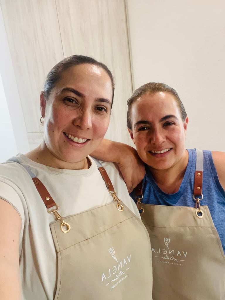 Two smiling women wearing matching tan aprons with brown straps standing together indoors.