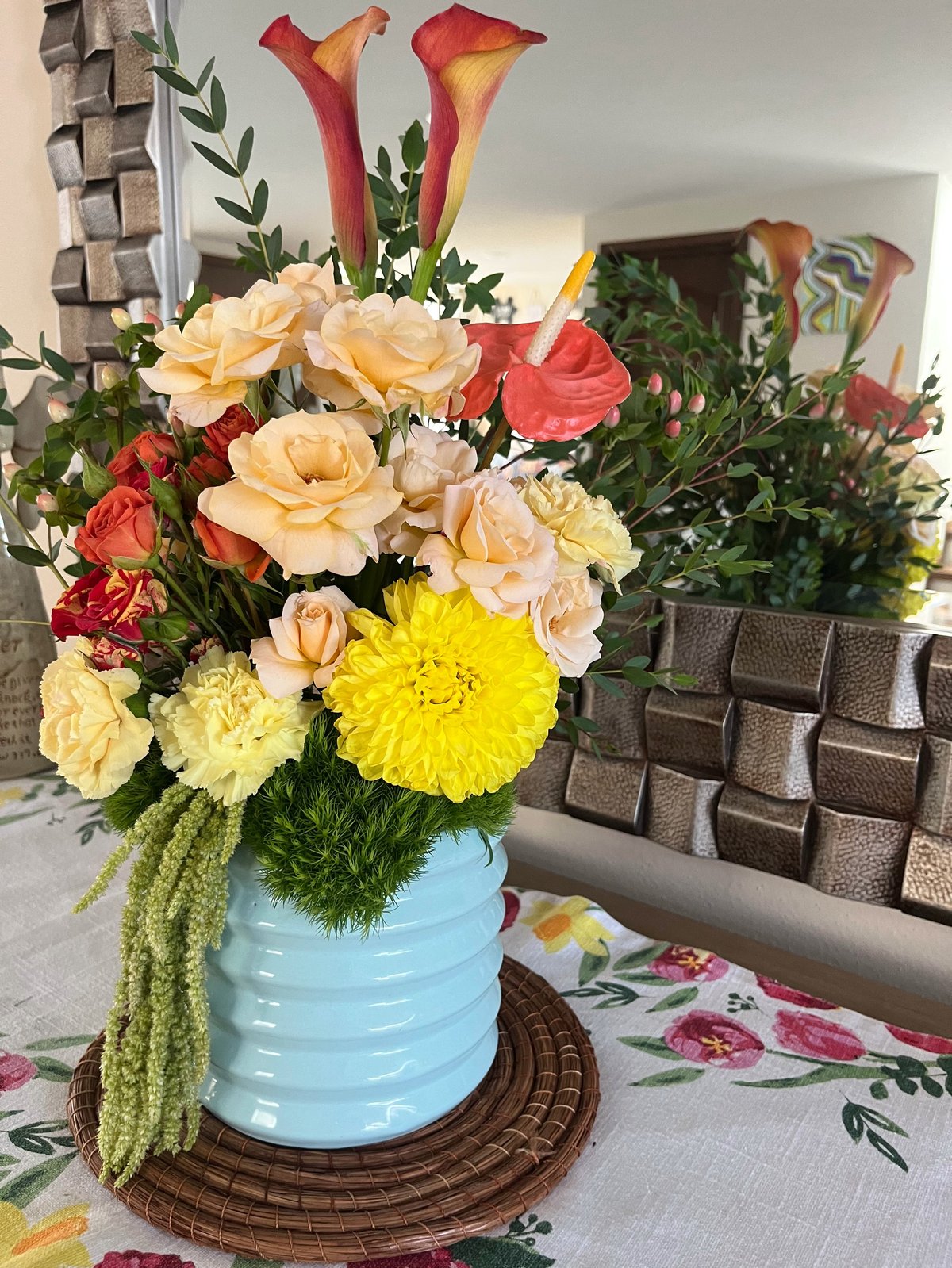 Vibrant floral arrangement in a light blue ceramic vase with peach roses, red calla lilies, yellow dahlias, and cascading green foliage on a woven coaster