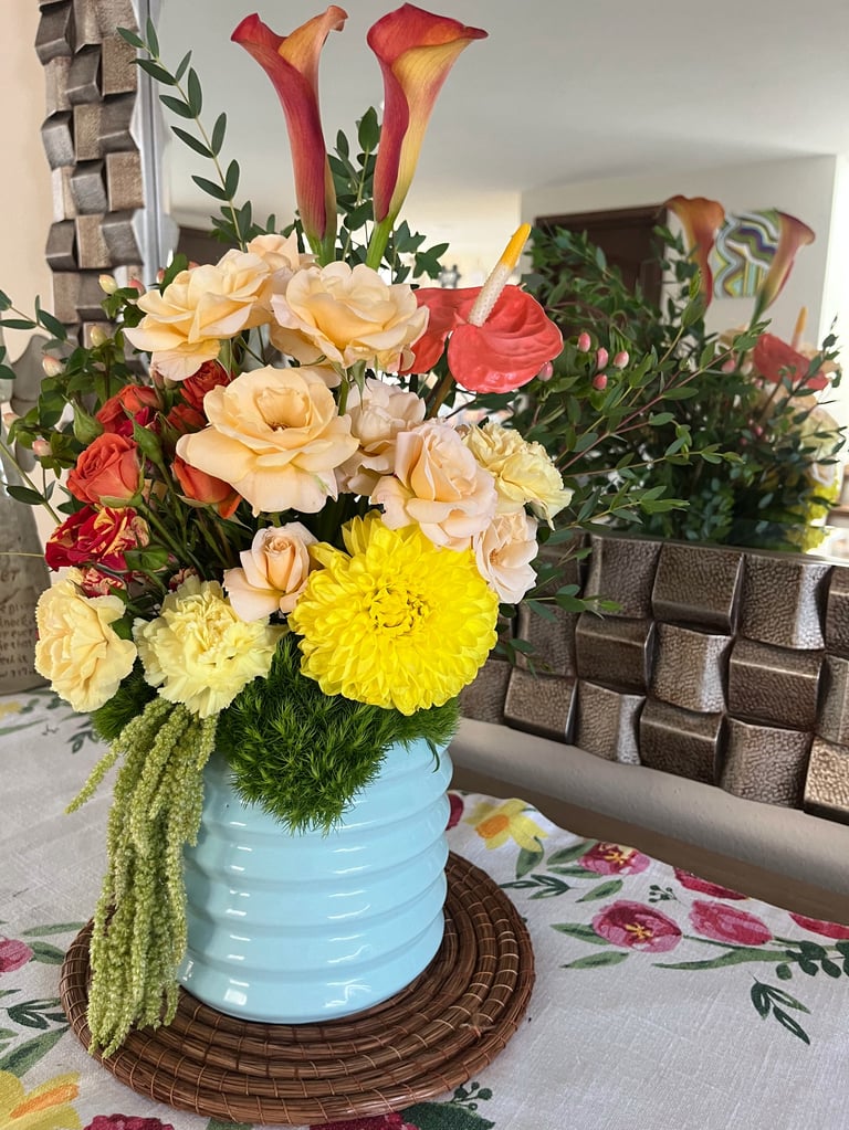 Vibrant floral arrangement in a light blue ceramic vase with peach roses, red calla lilies, yellow dahlias, and cascading green foliage on a woven coaster