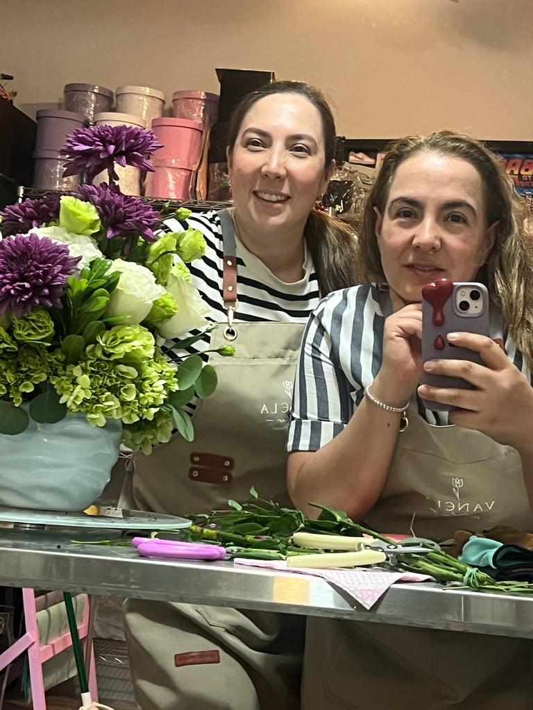 Two women taking a selfie at a florist workspace with purple and green flowers in a blue vase behind them