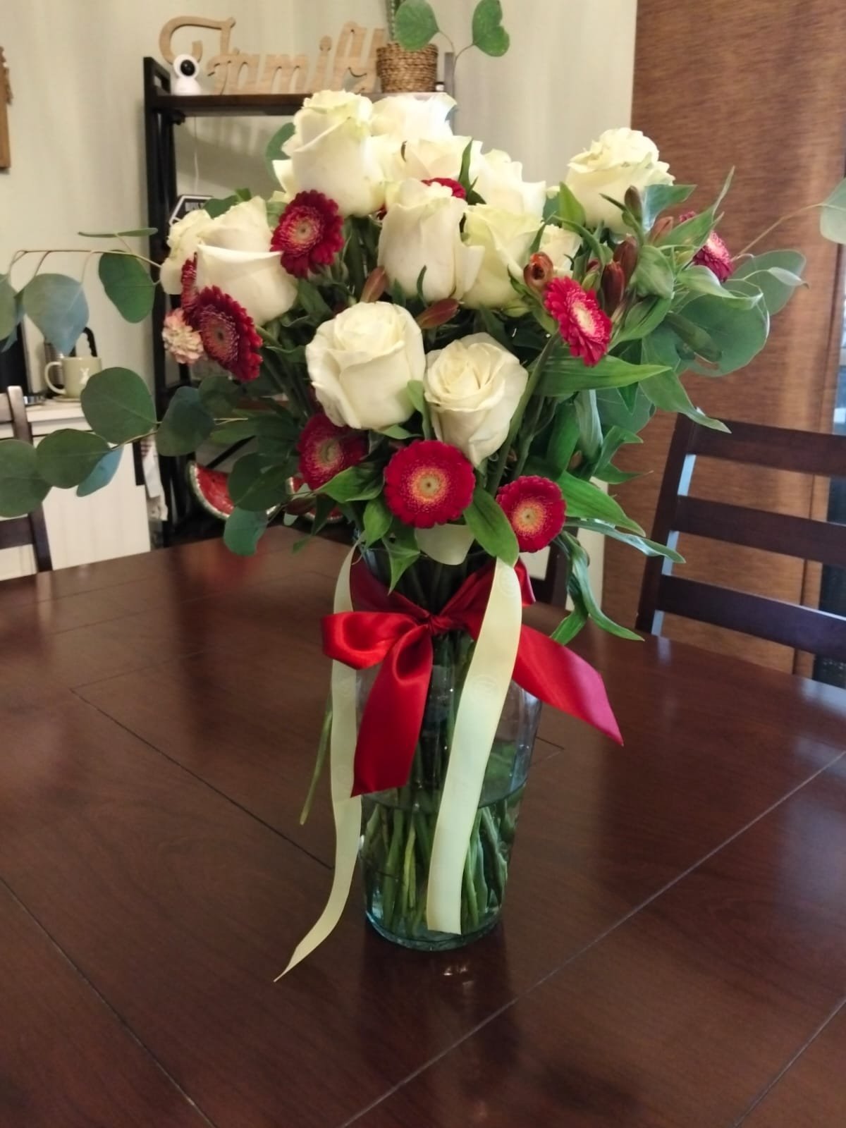 Floral bouquet in vase with white roses, red gerbera daisies, and eucalyptus greenery tied with red ribbon on dining table