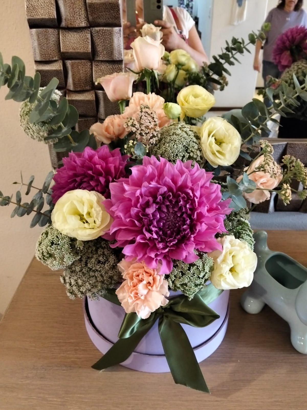 Colorful flower arrangement in a lavender round box with magenta dahlias, cream roses, peach carnations, white filler flowers, and eucalyptus greenery tied with a dark green ribbon.