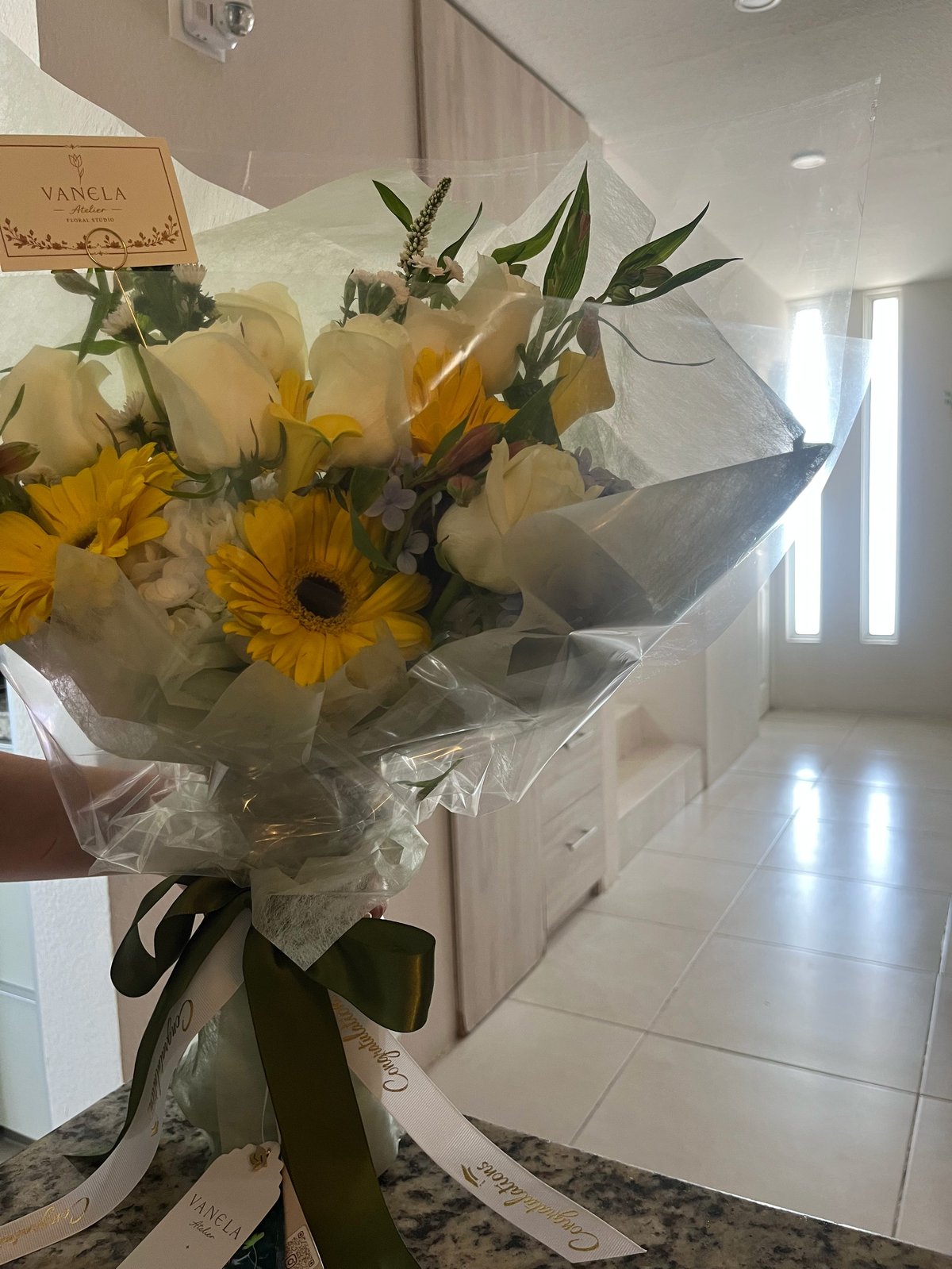 Bouquet of white roses and yellow gerbera daisies wrapped in clear cellophane with a black ribbon bow, displayed indoors