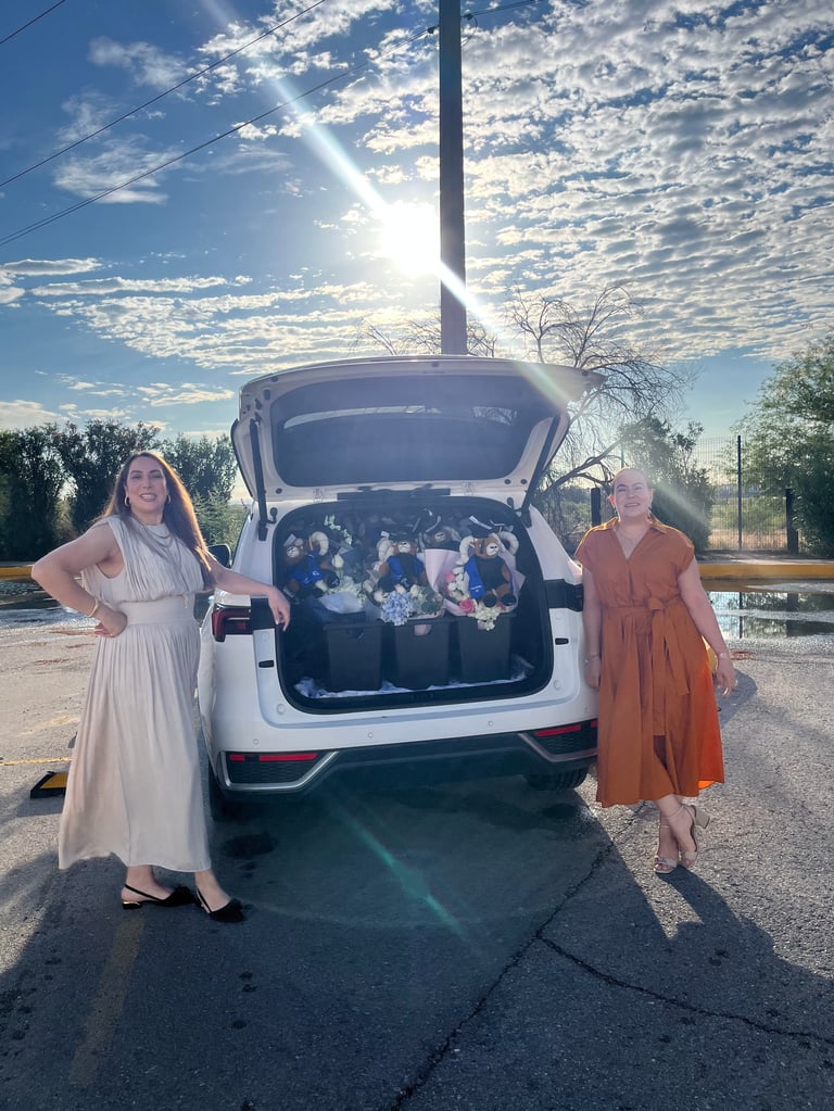 Two women standing beside a white car with its trunk open, displaying a large group of people inside against a bright sky backdrop.