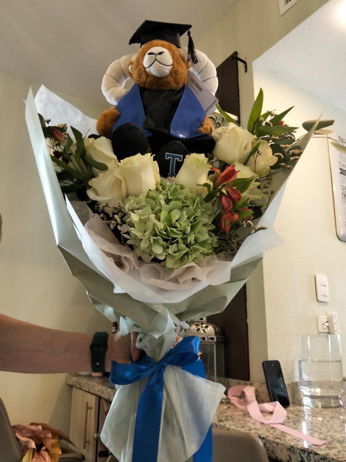 Graduation teddy bear wearing cap and gown positioned in a floral bouquet wrapped in white paper with blue ribbon
