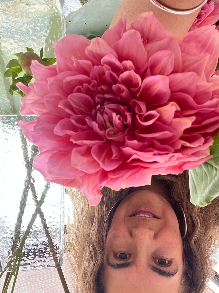 Woman holding a large pink dahlia flower above her head, smiling at camera in upside-down selfie view