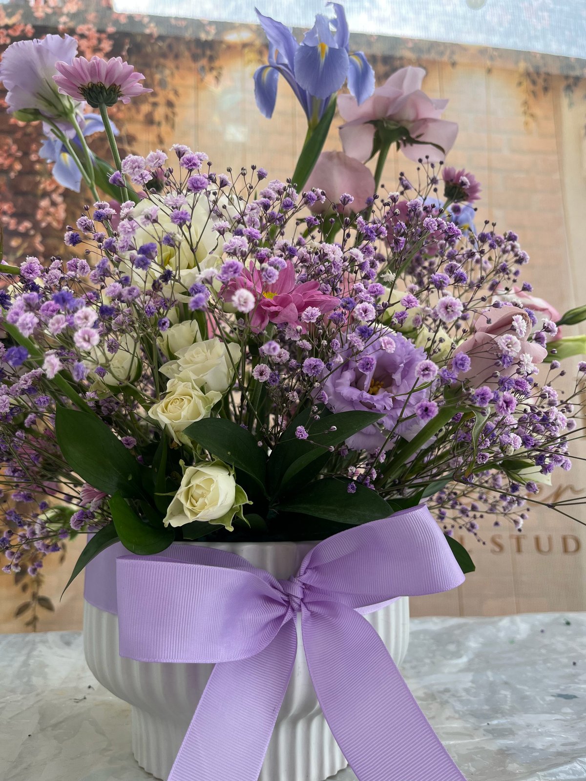 Floral arrangement in white vase with purple ribbon bow, featuring purple statice, white roses, pink carnations, and blue iris flowers