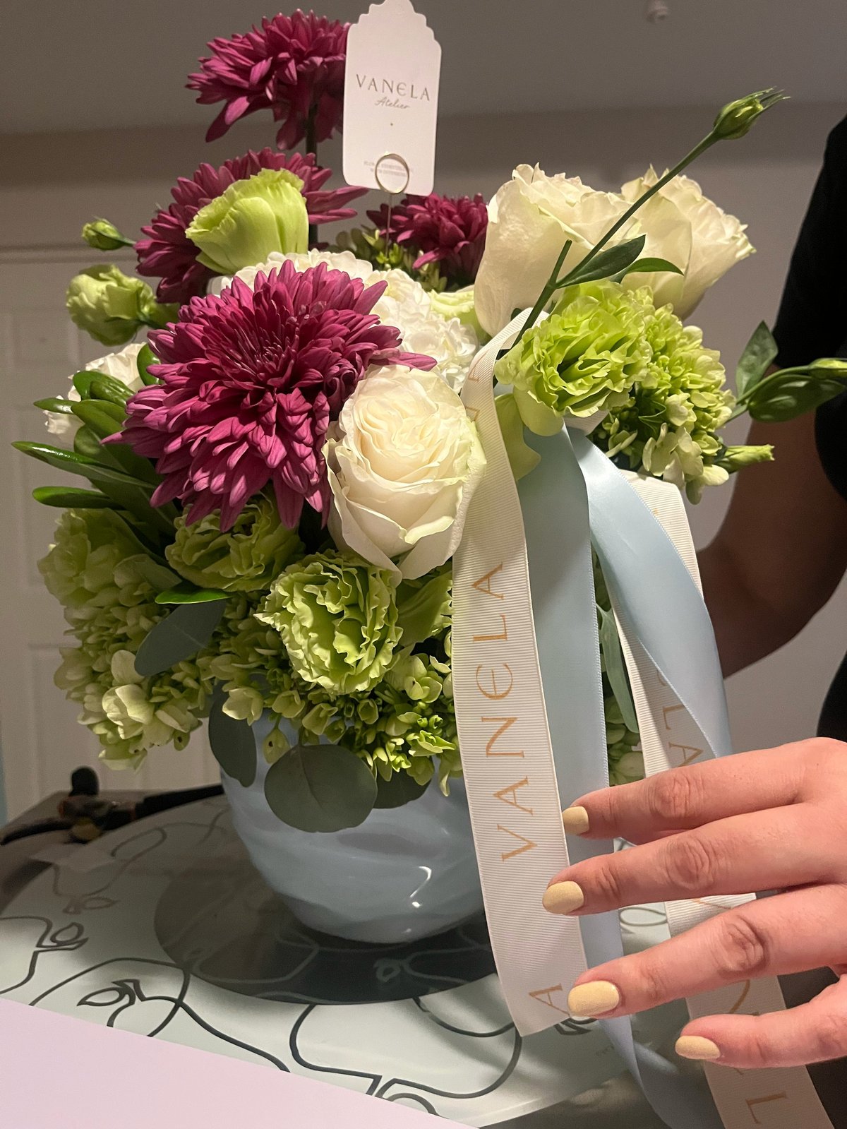 Hand holding white ribbon attached to white vase arrangement of magenta dahlias, white roses, and green carnations with VANELA branding