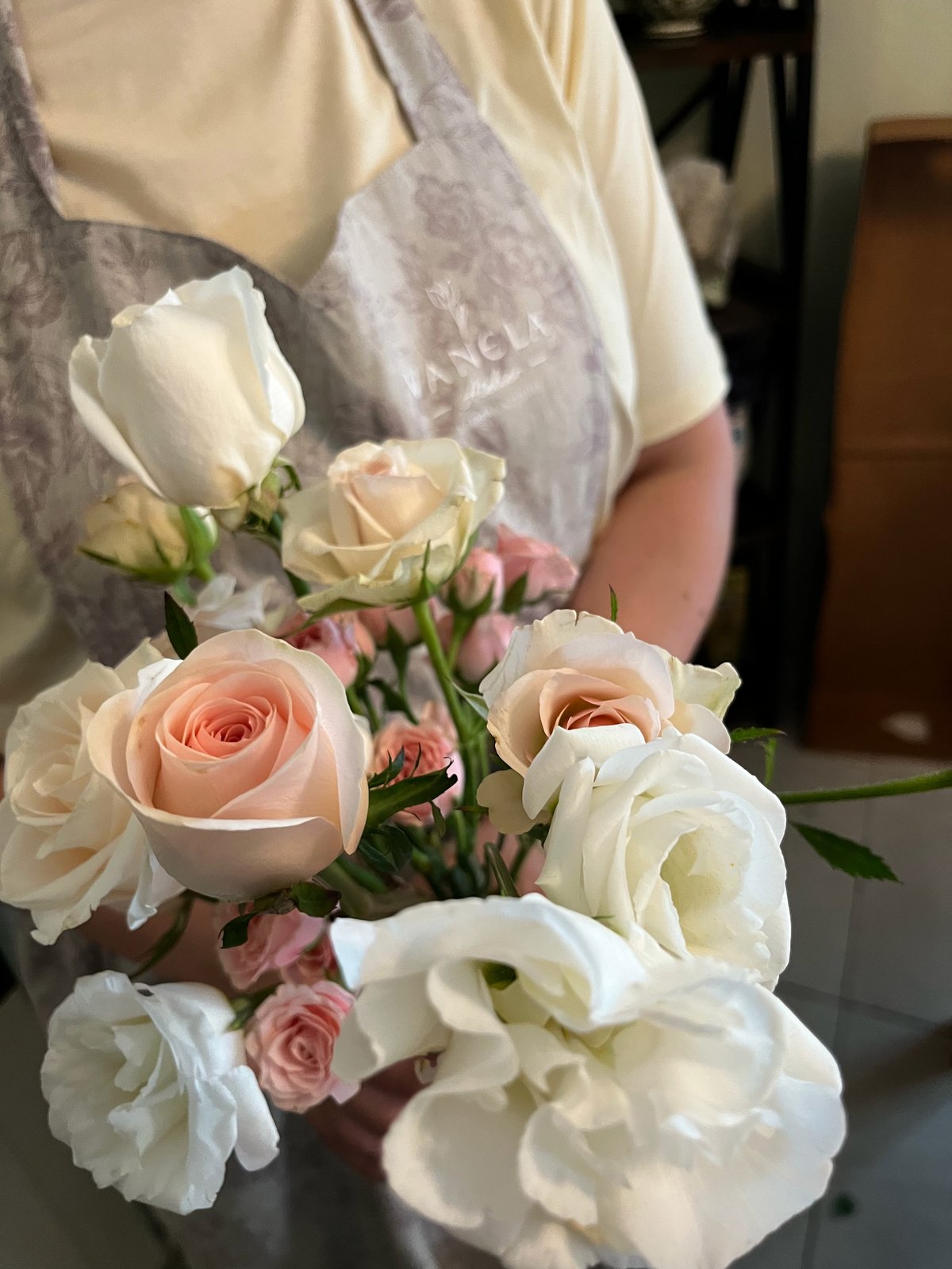 Person in gray apron holding a bouquet of white, peach, and pink roses with green stems and foliage