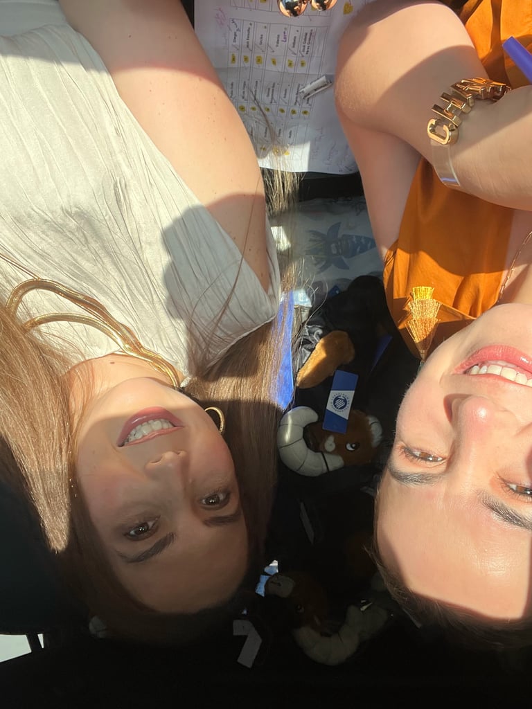 Two people posing upside down for a selfie with colorful eyeshadow and gold jewelry, sunlit indoor setting