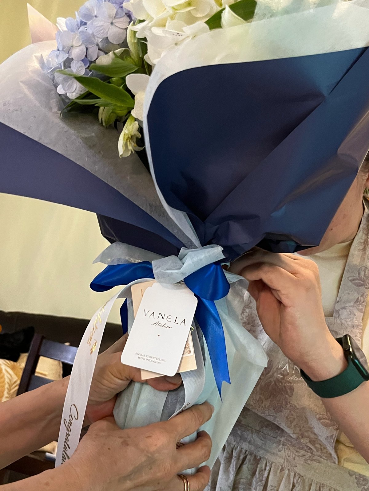 Hands holding an elegant bouquet of blue and white hydrangeas wrapped in navy blue paper with blue ribbon and a Vanela tag