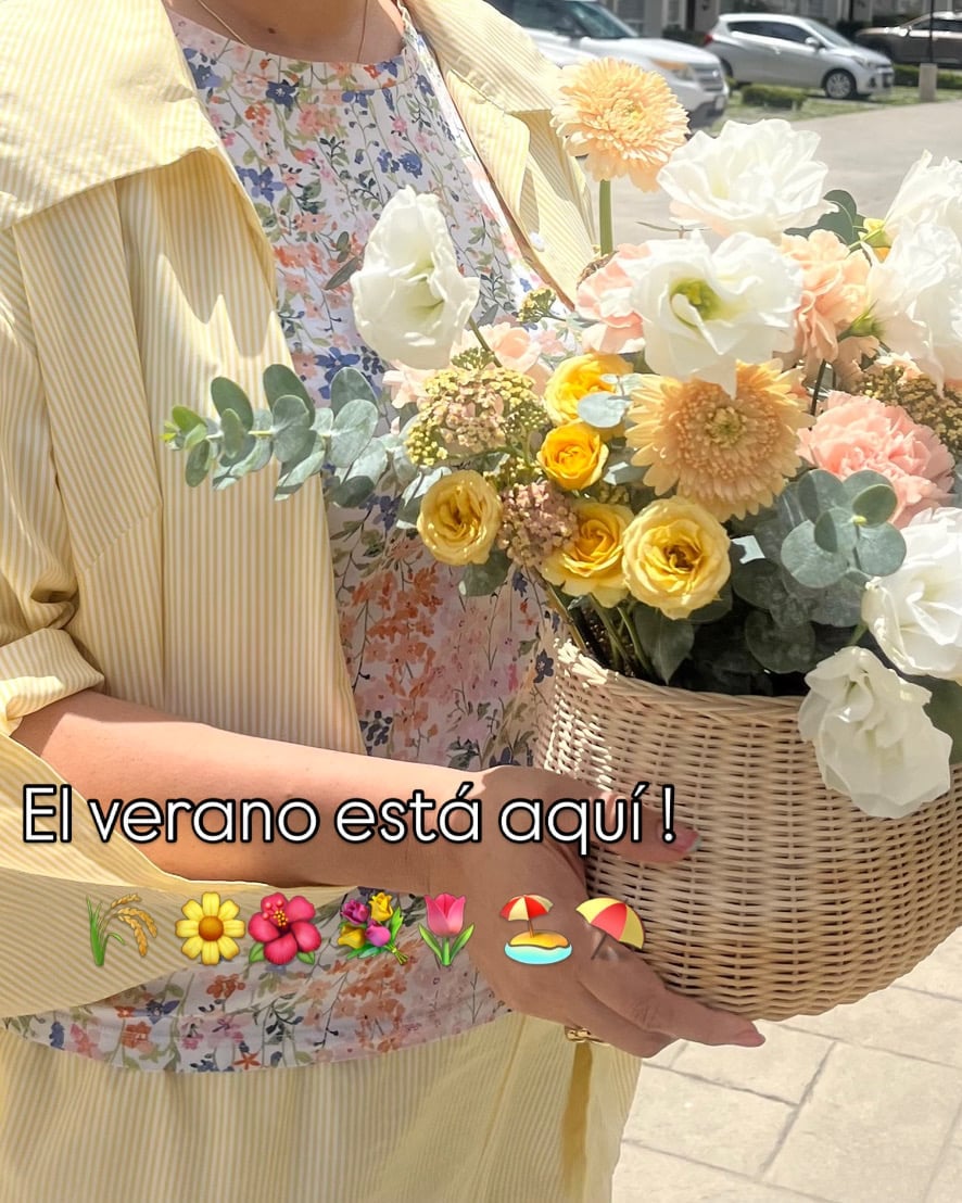 Person holding a wicker basket of colorful flowers including yellow roses, white peonies, and greenery in summer sunlight