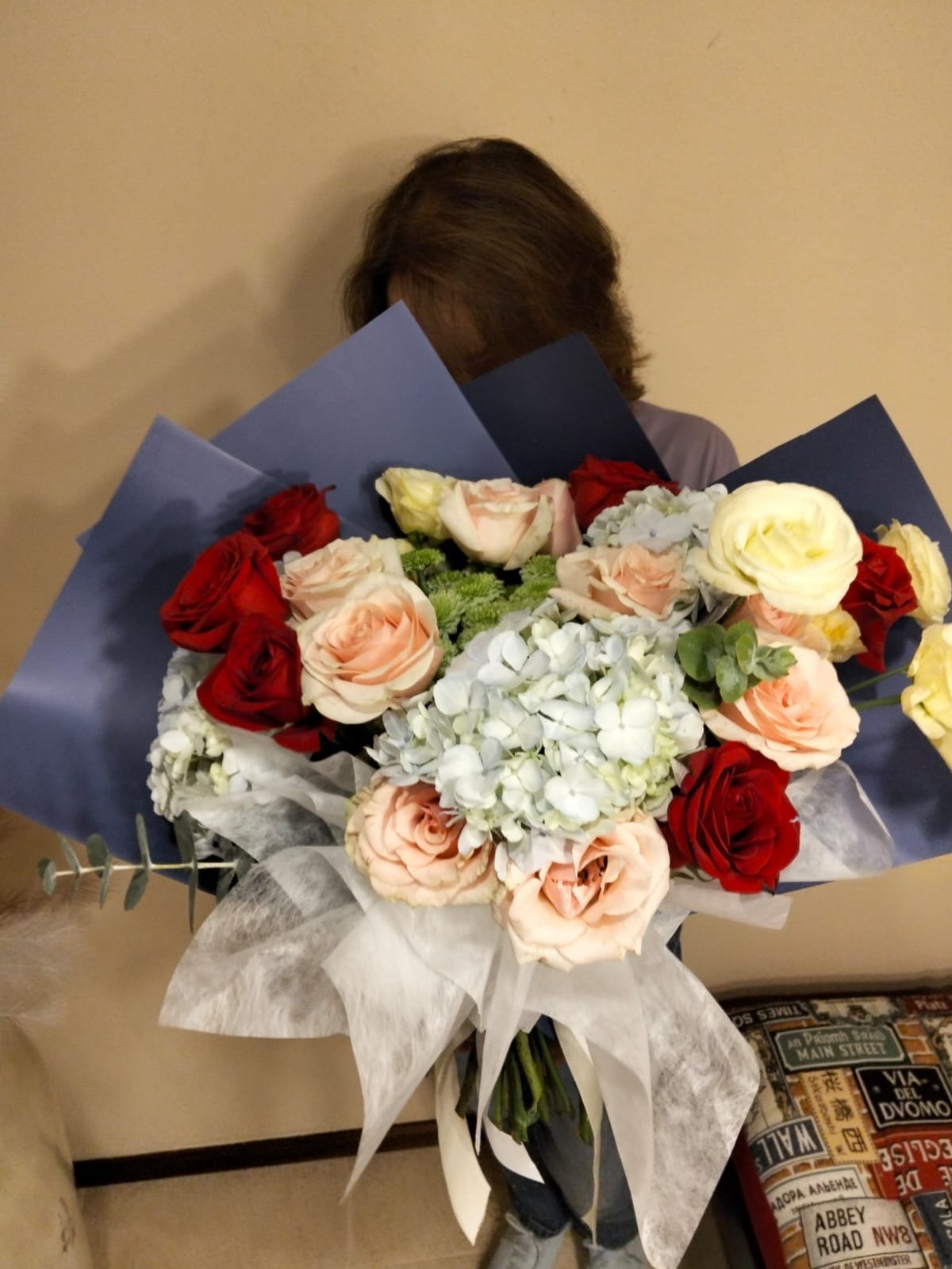 Person holding a large bouquet of red roses, peach and yellow flowers with white hydrangeas wrapped in blue paper and white ribbon