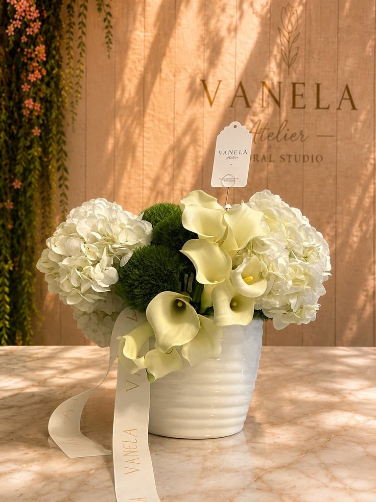 Arreglo elegante de flores blancas en maceta de cerámica con calas crema y hortensias blancas sobre mostrador de mármol, ideal para condolencias