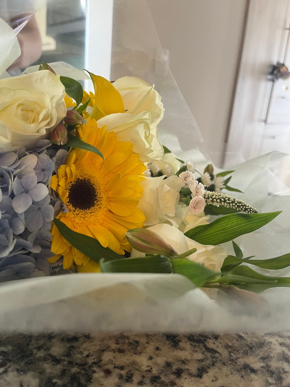 Bright floral bouquet with yellow gerbera daisy and cream roses arranged with green foliage in a white vase on a granite countertop