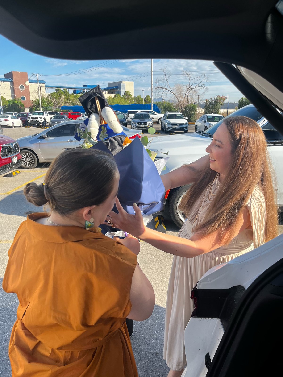 Two women in a parking lot, one wearing orange and one in white, holding up a black graduation cap near a vehicle