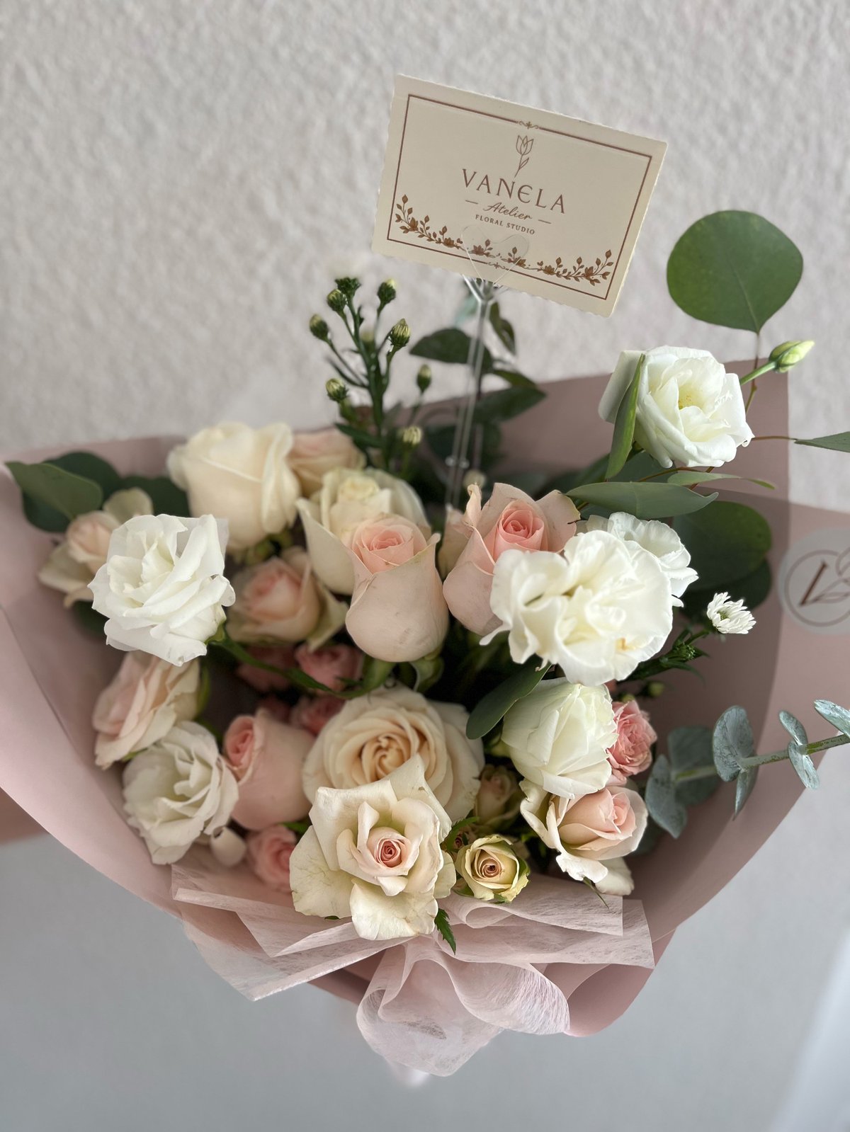 Elegant bouquet of white, cream, and blush roses with green foliage wrapped in pink tissue paper, featuring a Vanela branded card