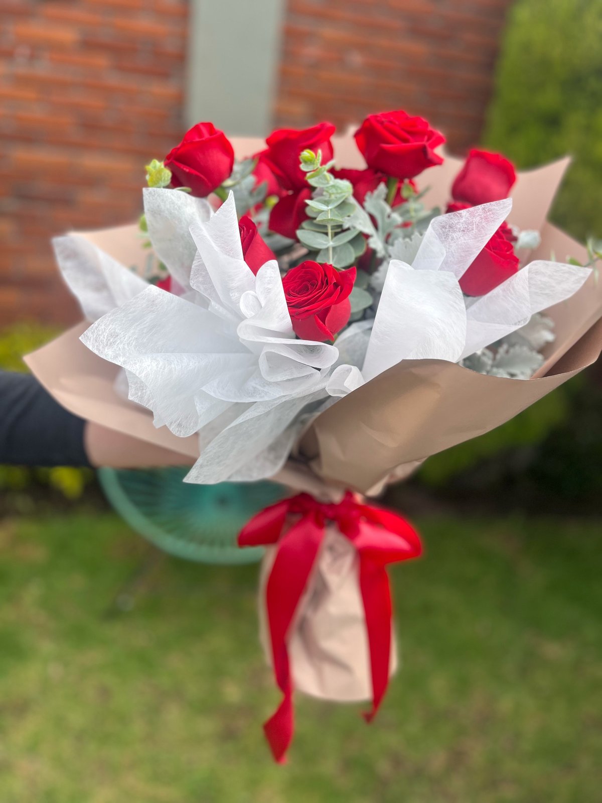 Bouquet of red roses wrapped in white tissue paper and kraft paper with red ribbon bow, held outdoors against a brick building