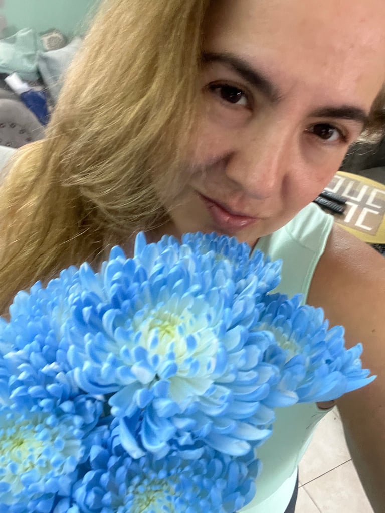 Woman with blonde hair holding a large blue chrysanthemum flower, smiling at camera in indoor setting