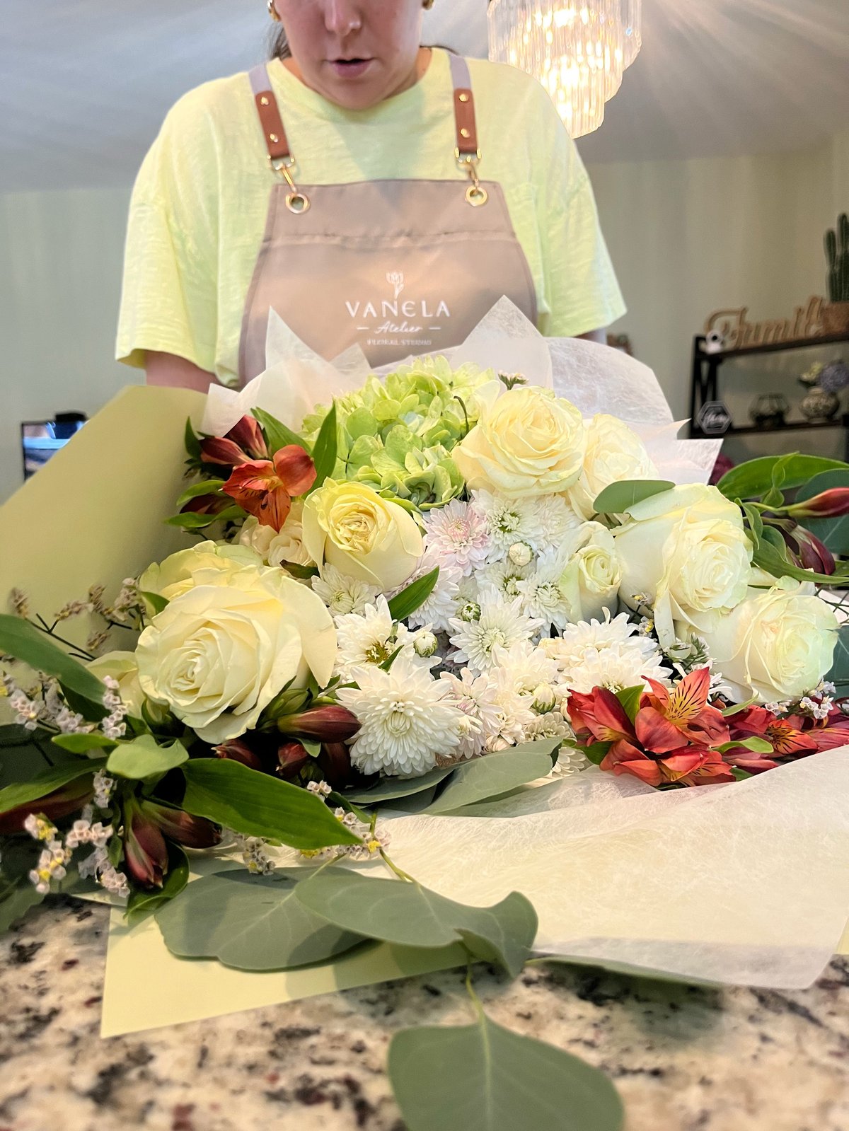Florist in yellow apron arranging a bouquet with cream roses, red flowers, and white blooms on a work table