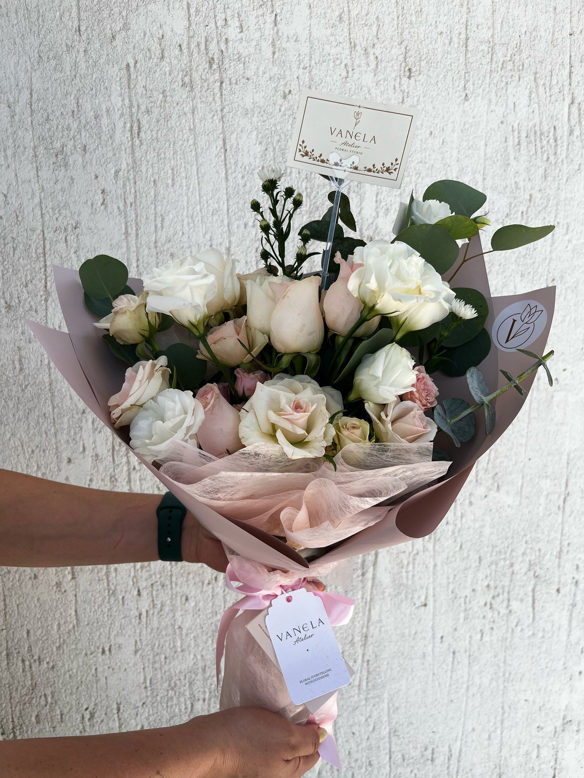 Person holding an elegant bouquet of cream and pink roses with green foliage and black dried flowers, wrapped in pink tissue paper with Vanela brand tags