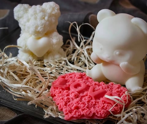 Handmade soap figurines displayed on straw: cream-colored teddy bear, white pig, and pink rose-shaped soaps