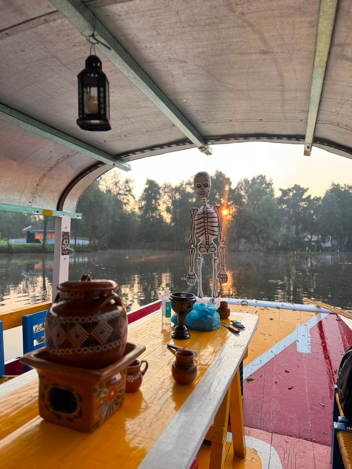 Interior de una trajinera con decoración de esqueleto, linterna y mesa de madera junto al canal al atardecer
