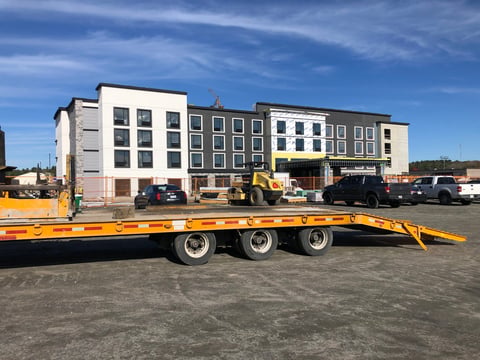 Yellow flatbed trailer parked in lot with multi-story residential buildings and construction vehicles in background
