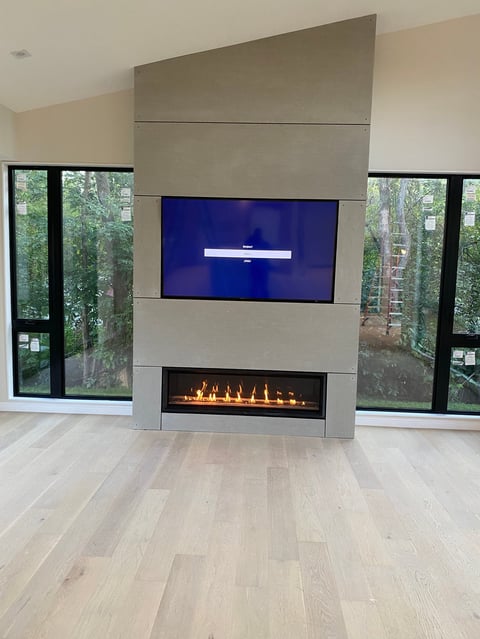 Modern fireplace with blue TV mounted above, surrounded by tall windows overlooking trees in a bright living room with light wood flooring.