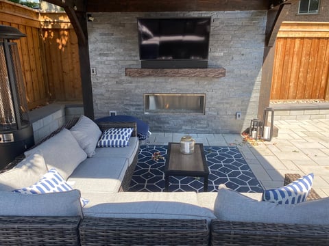 Modern outdoor patio with gray seating, blue and white pillows, geometric rug, TV mounted on brick wall, and metal fireplace insert