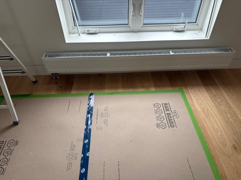 Construction flooring with underlayment, green tape, and blue leveling tools under a ceiling window and baseboard heater