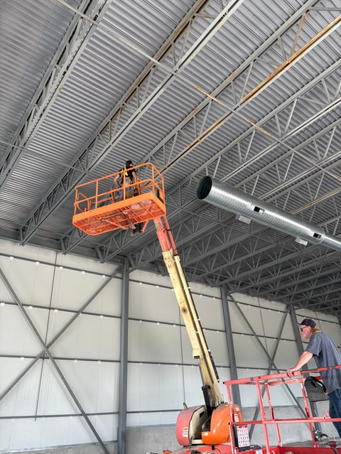 Worker operating an orange articulating boom lift inside a warehouse with corrugated metal roof and white walls