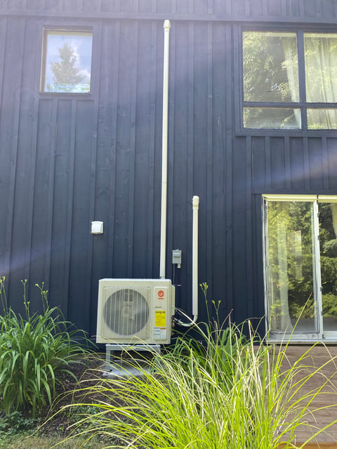 Heat pump unit mounted on black corrugated metal building with windows and ornamental grass in foreground