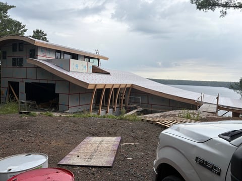 Modern house under construction on lakeside property with curved wooden supports, partially parked vehicle in foreground