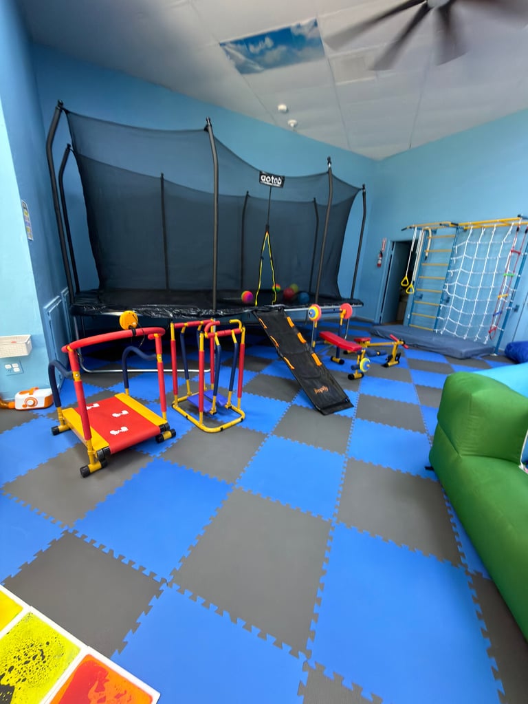 Indoor play area with colorful foam mat flooring, trampoline, and various childrens gym equipment including climbing structures and obstacles.