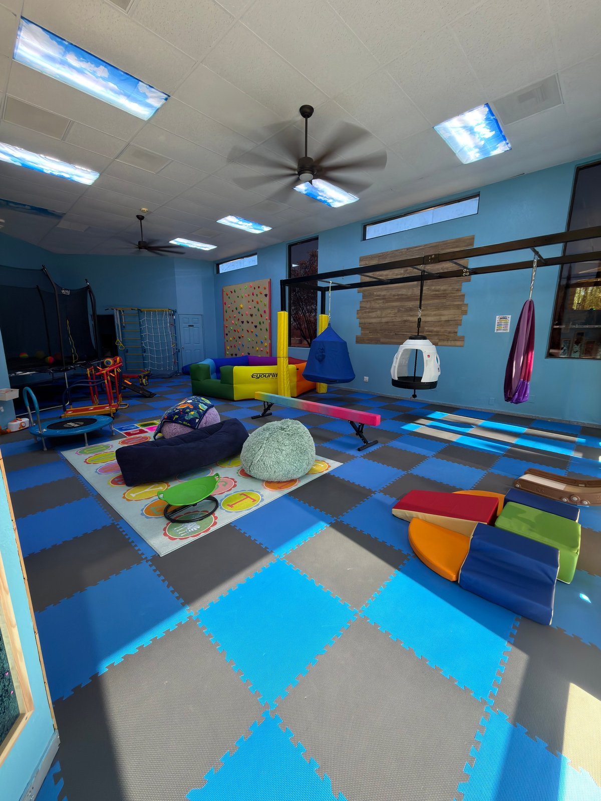Colorful indoor childrens play gym with blue walls, checkered floor, climbing equipment, swings, foam blocks, and various exercise apparatus