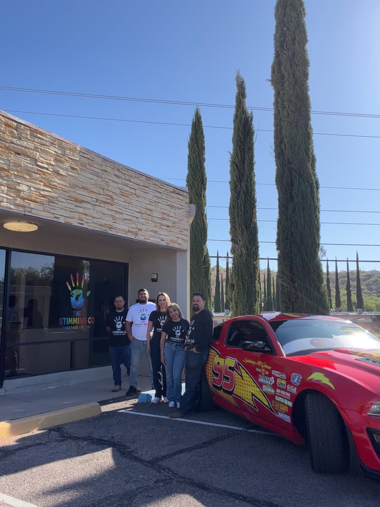 Group of six people standing next to a red custom hot rod car outside a building with tall cypress trees in background