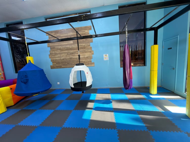 Colorful indoor play area with hanging equipment, foam padding, blue checkered floor, and various play structures including a blue cone and suspended gear