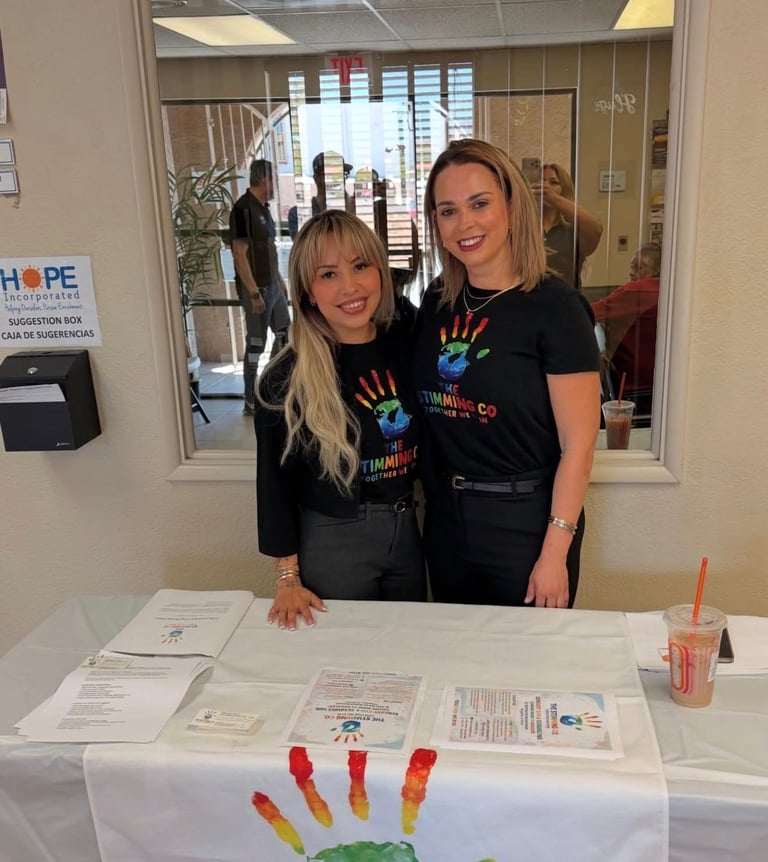 Two women in black shirts smiling at a charity table with documents and a rainbow handprint craft decoration