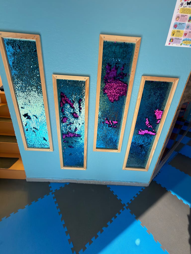 Four framed liquid sensory boards with moving glitter and colorful beads mounted on light blue wall in playroom