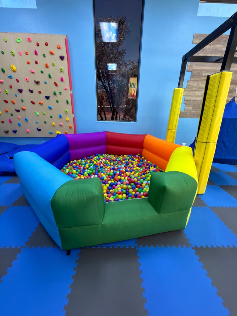 Colorful indoor play area with green foam couch filled with multicolored ball pit, climbing wall, and various foam equipment