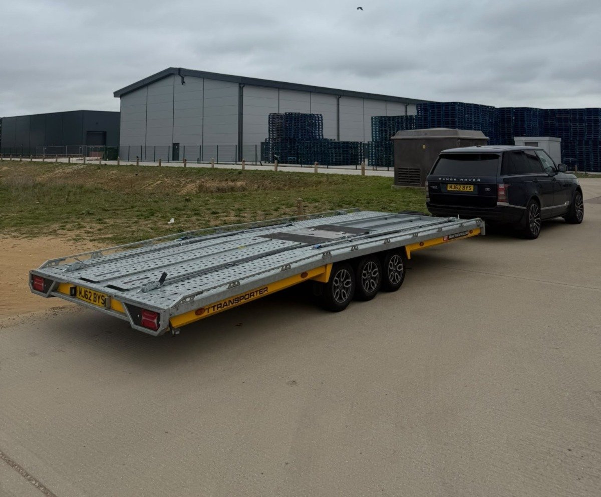 A black SUV towing an empty yellow and gray flatbed trailer in a parking lot with industrial warehouses in the background.
