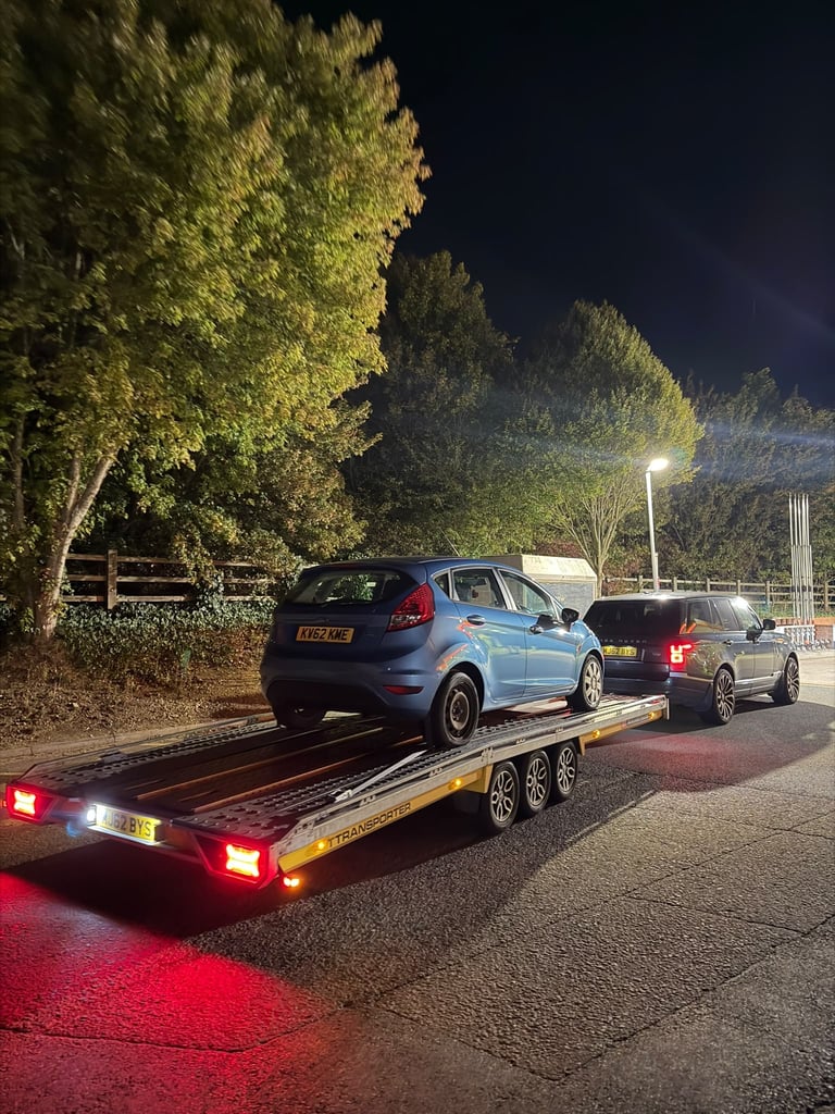 Blue car being loaded onto a flatbed tow truck at night in a rural area with illuminated trees